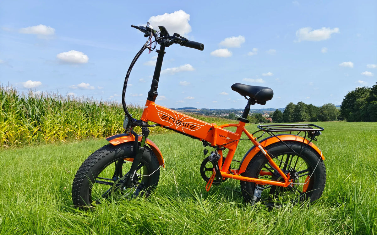 Vélo électrique pliant ENGWE EP-2 Pro orange à pneus larges et porte-bagages dans les hautes herbes près d’un champ de maïs