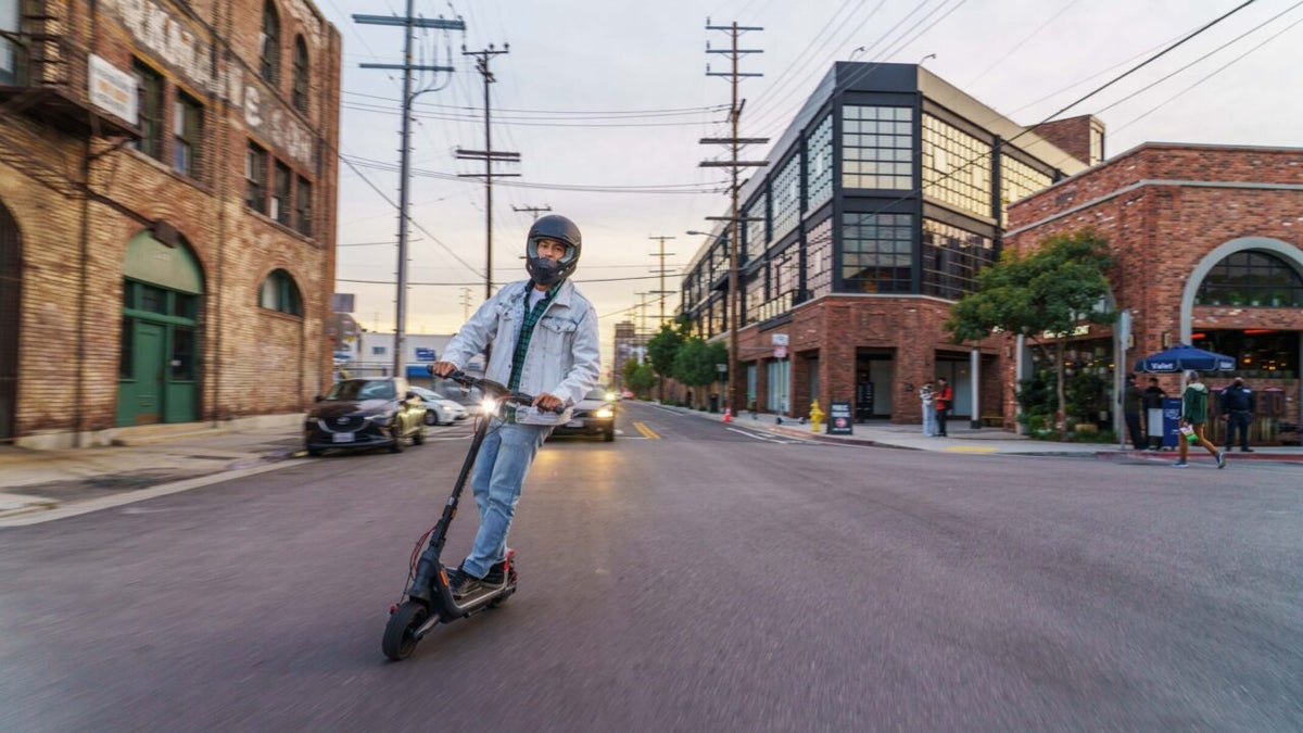 Homme avec un casque intégral noir sur une trottinette électrique, phare allumé, dans une rue au crépuscule