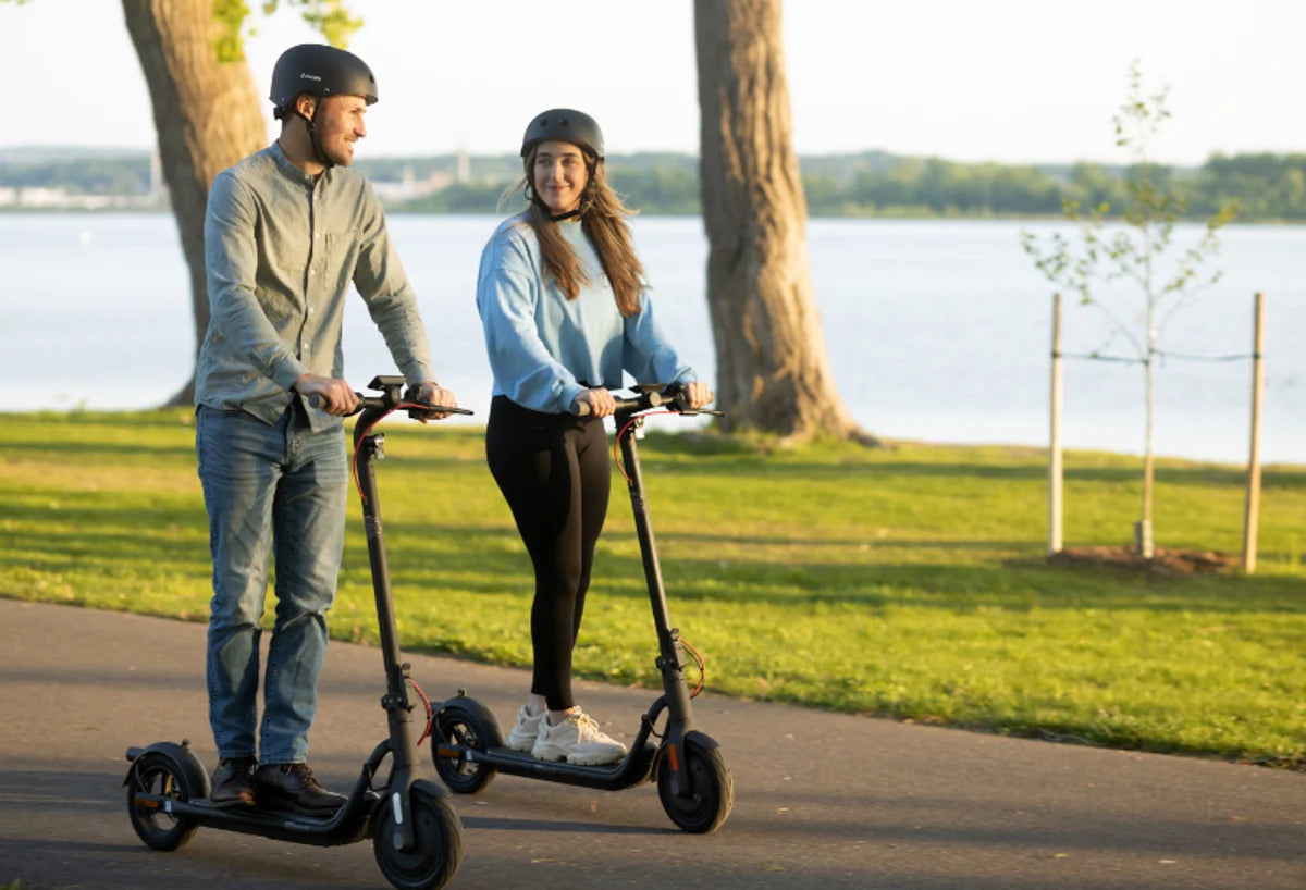Deux personnes casquées roulent en trottinette électrique NAVEE sur un chemin au bord d’un lac