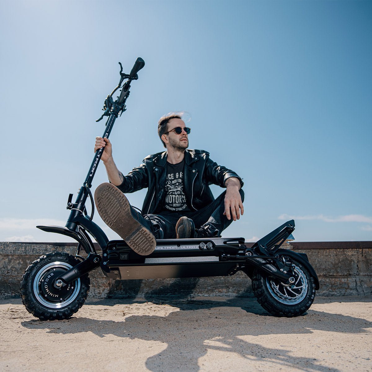 Homme en blouson de cuir et lunettes de soleil assis sur une Dualtron Ultra noire aux pneus tout-terrain, sur le sable