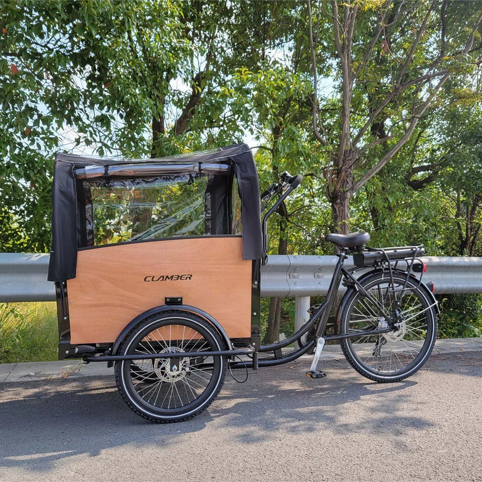 Triporteur avec caisse en bois et capote noire à fenêtres, garé sur une route près d’une glissière, arbres verts derrière