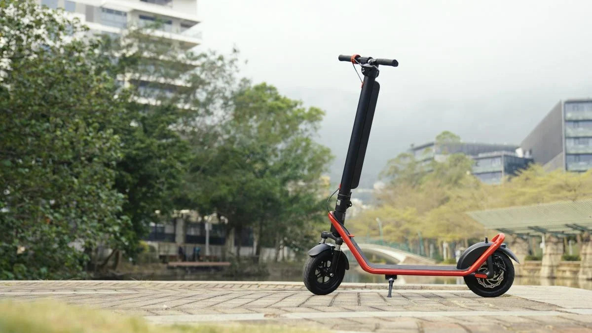 Trottinette électrique HX noire à cadre rouge sur sa béquille, sur une promenade pavée, arbres et immeubles modernes derrière
