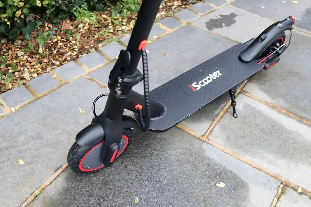 Trottinette électrique iScooter noire aux jantes liserées de rouge, sur sa béquille, sur des dalles grises humides