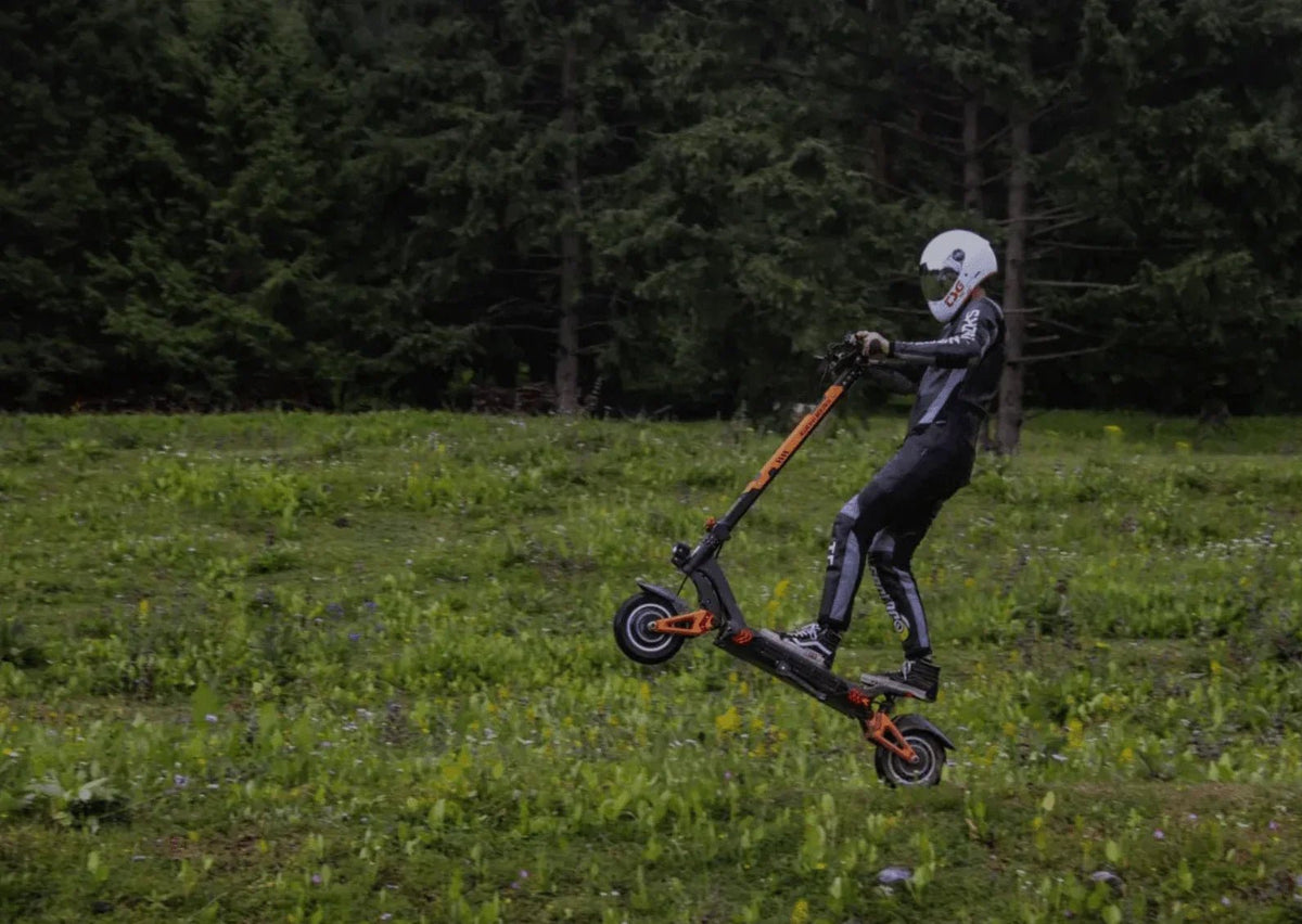 Pilote casqué en combinaison sautant avec une trottinette noire et orange au-dessus d’un pré fleuri, devant des sapins