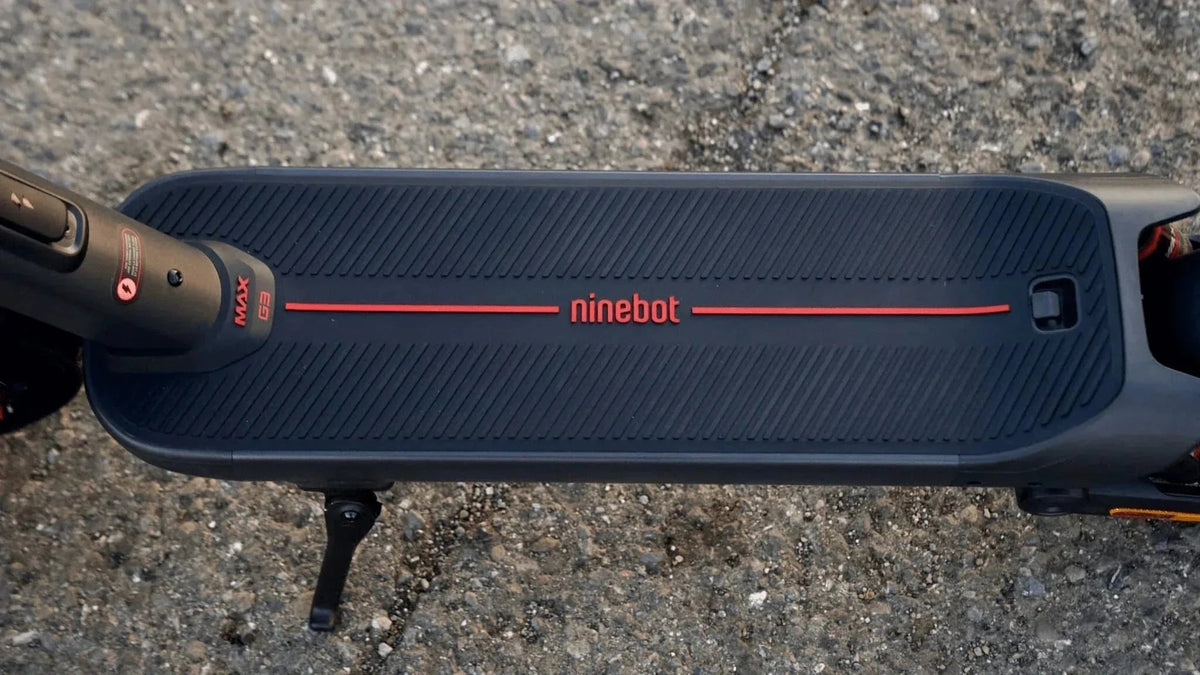 Vue de dessus du plateau d’une Ninebot MAX G3 noire, logo ninebot rouge et béquille dépliée, sur un bitume gravillonné