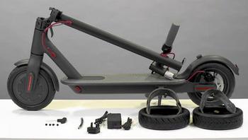 Trottinette électrique gris foncé pliée, détails rouges, sur table blanche, devant deux pneus avec chambre et un chargeur