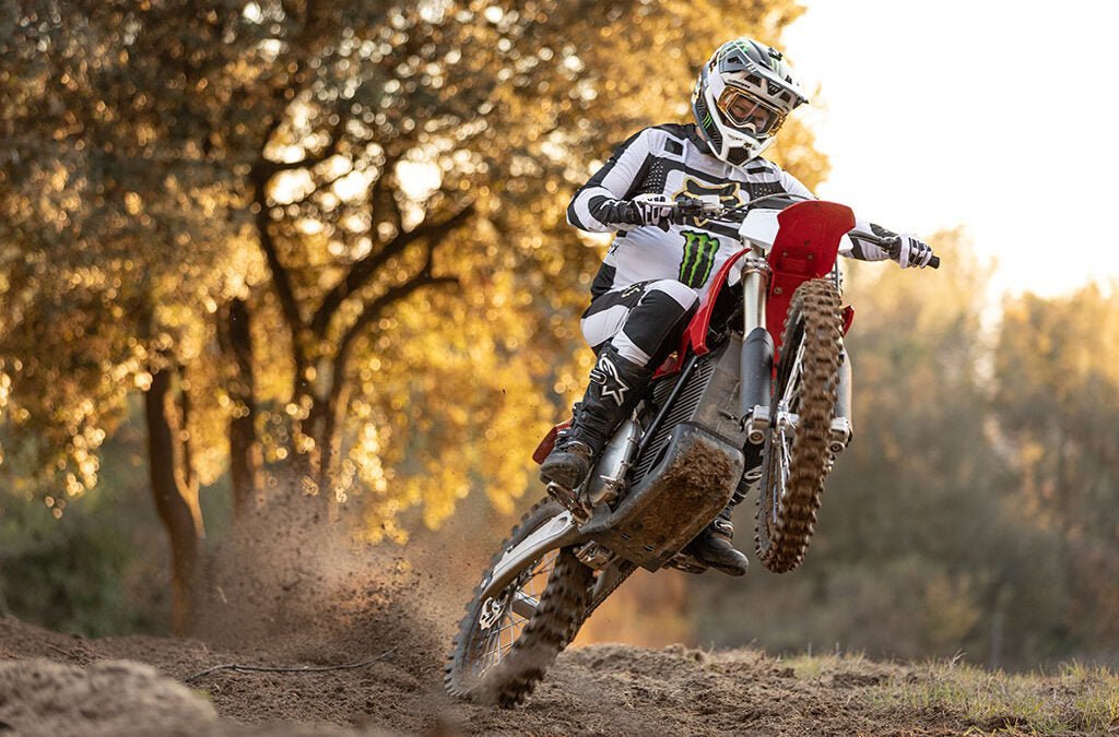 Pilote en tenue blanche et noire qui lève la roue avant d’une moto cross rouge sur piste en terre, arbres en contre-jour