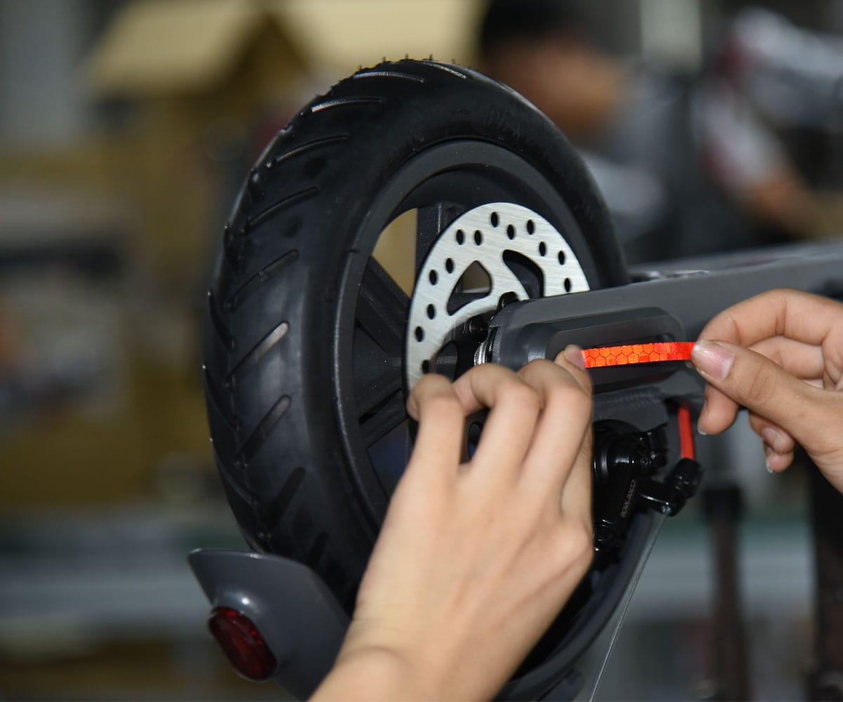 Des mains posent une bande réfléchissante rouge sur la fourche arrière d’une trottinette électrique, près du disque de frein