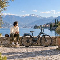Vélo électrique OneSport OT12 Pro sur un quai pavé au bord d'un lac, avec un homme assis sur le muret à côté et des montagnes en arrière-plan