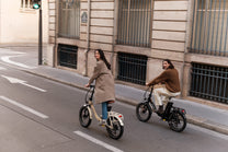 Deux personnes à vélo dans une rue en ville, chacune sur un vélo pliant, l'un beige et l'autre noir