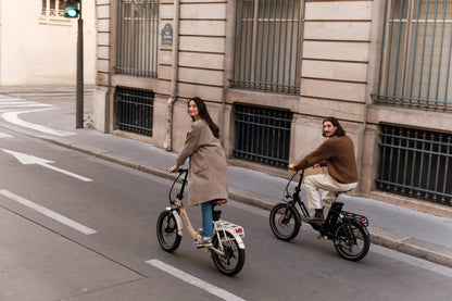 Deux personnes à vélo dans une rue en ville, chacune sur un vélo pliant, l'un beige et l'autre noir