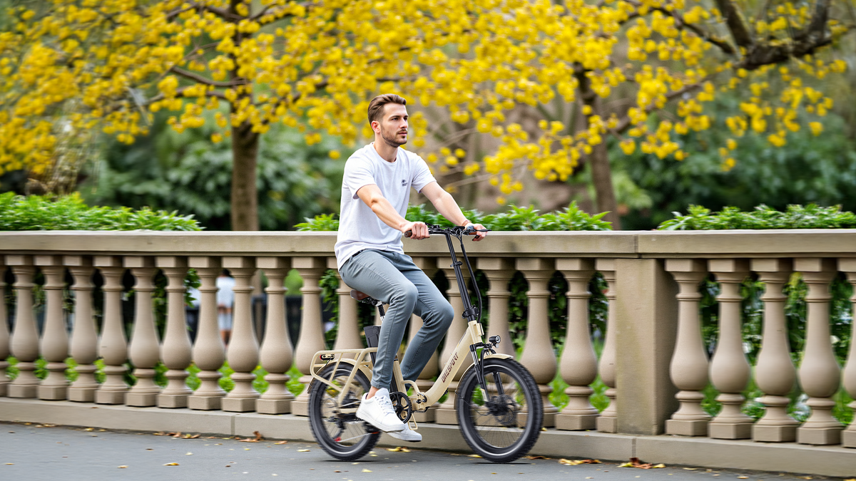 Homme en t-shirt blanc à vélo sur un OneSport OT16 Pro beige devant une balustrade en pierre et des arbres en fleurs jaunes