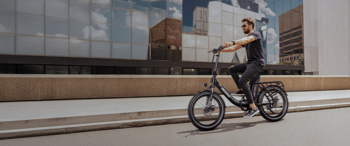 Homme avec des lunettes de soleil à vélo sur un OneSport OT16 Pro le long d'une rue devant une façade vitrée