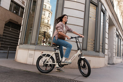Femme à vélo sur l'OneSport OT16 Pro, vélo électrique blanc vu de côté dans une rue en ville près d'un bâtiment en pierre