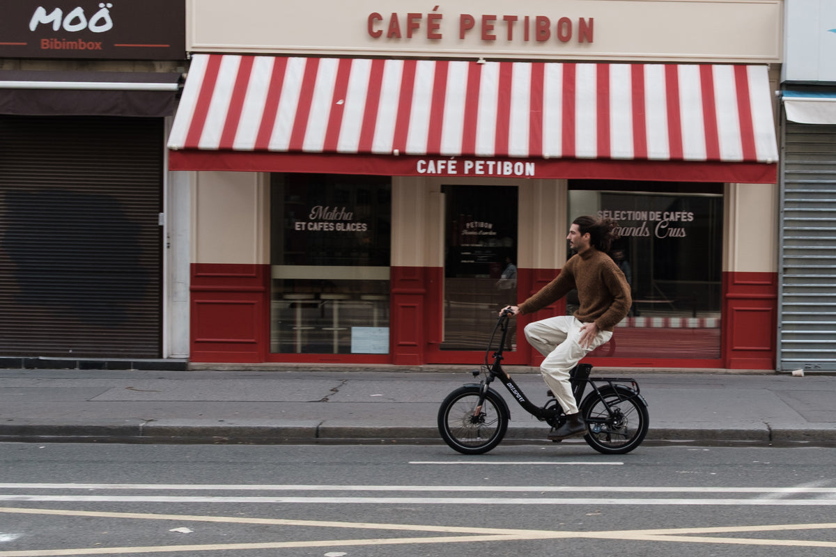 Homme à vélo sur l'OneSport OT16 Pro, vélo noir, vue de côté devant un café avec un store à rayures rouges et blanches