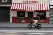 Homme à vélo sur l'OneSport OT16 Pro, vélo noir, vue de côté devant un café avec un store à rayures rouges et blanches