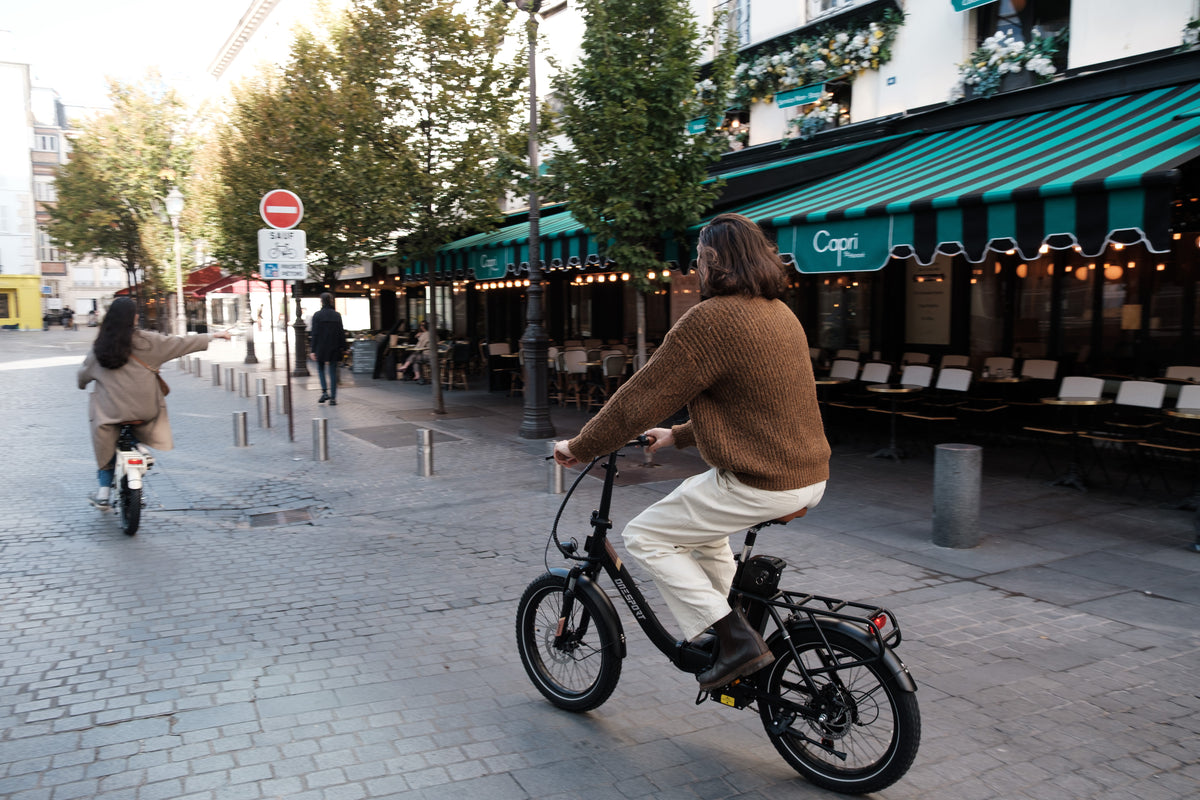 Personne à vélo sur l'OneSport OT16 Pro, vélo noir, dans une rue pavée devant un café avec un store à rayures vertes