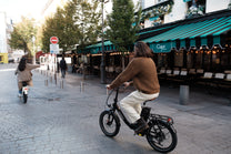 Personne à vélo sur l'OneSport OT16 Pro, vélo noir, dans une rue pavée devant un café avec un store à rayures vertes