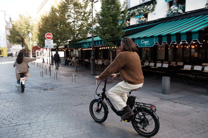 Personne à vélo sur l'OneSport OT16 Pro, vélo noir, dans une rue pavée devant un café avec un store à rayures vertes