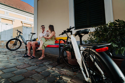 OneSport WT05, vélo blanc au premier plan, un couple assis sur un banc coloré et un autre vélo derrière sur une rue pavée