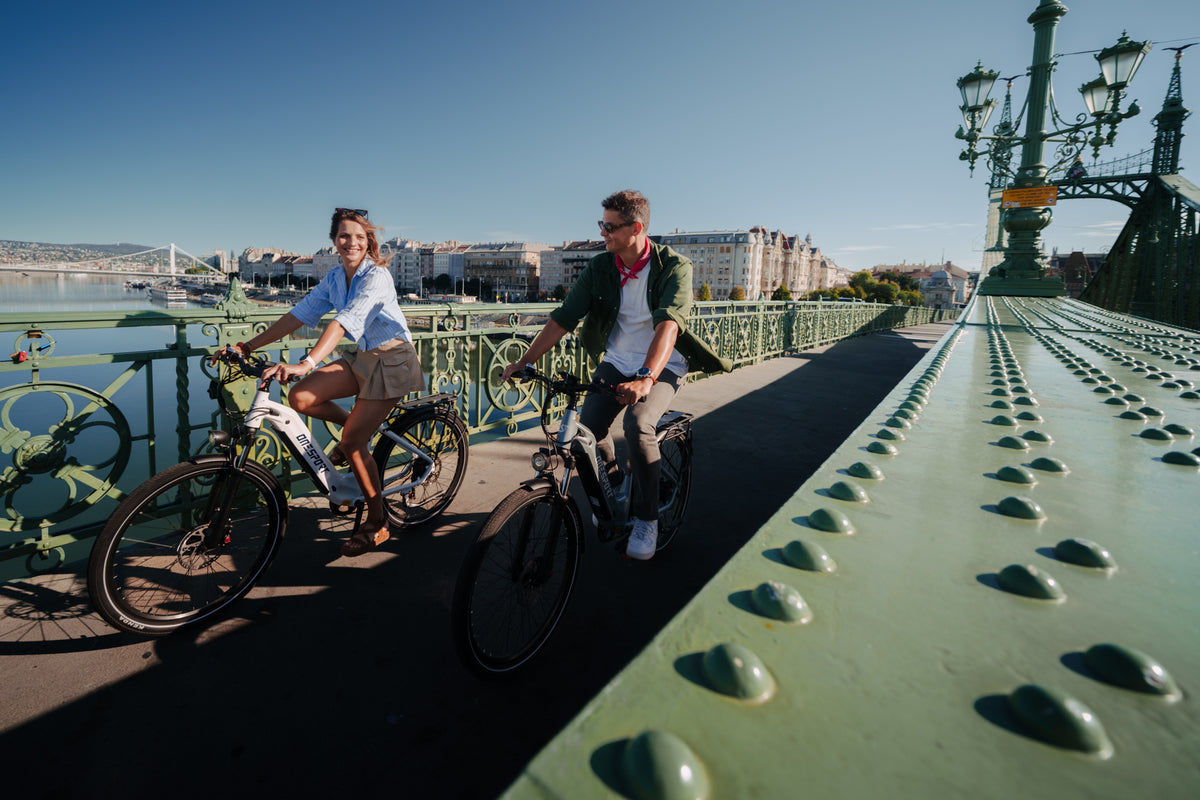 Deux personnes à vélo sur un pont vert au soleil avec des bâtiments et de l'eau à l'arrière-plan