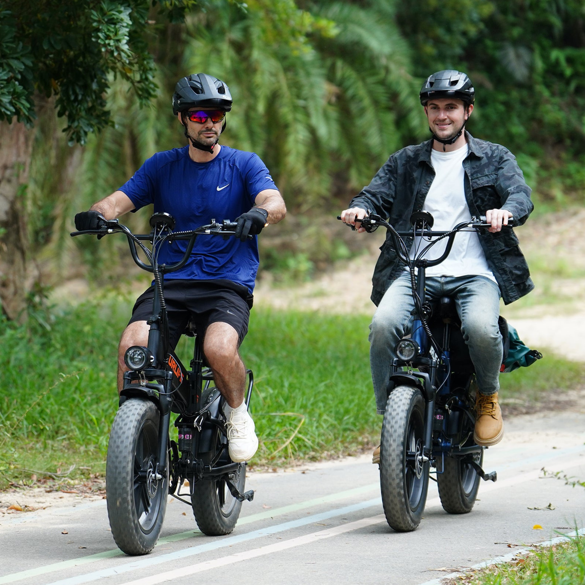Deux personnes casquées roulent côte à côte sur des vélos à pneus larges sur une route goudronnée dans la verdure