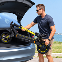 Homme portant des lunettes de soleil soulevant une trottinette électrique noire et jaune pliée avec le texte FREEBOY devant le coffre ouvert d'une voiture grise